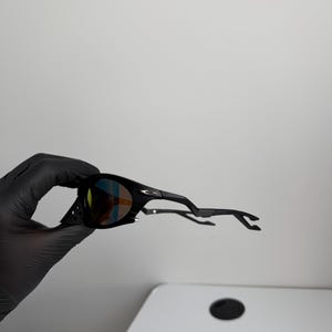 May include: Black sunglasses with a curved frame and tinted lenses. The lenses have a multi-colored reflective coating. The sunglasses are held by a gloved hand against a white background. The arms of the sunglasses are extended.