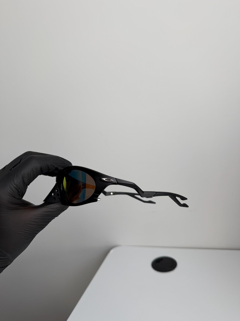 May include: Black sunglasses with a curved frame and tinted lenses. The lenses have a multi-colored reflective coating. The sunglasses are held by a gloved hand against a white background. The arms of the sunglasses are extended.