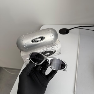 Puede incluir: Gafas de sol plateadas con lentes oscuras, presentadas con un estuche plateado a juego y una caja. Las gafas tienen un dise&ntilde;o elegante y moderno. El estuche presenta el logotipo de la marca.