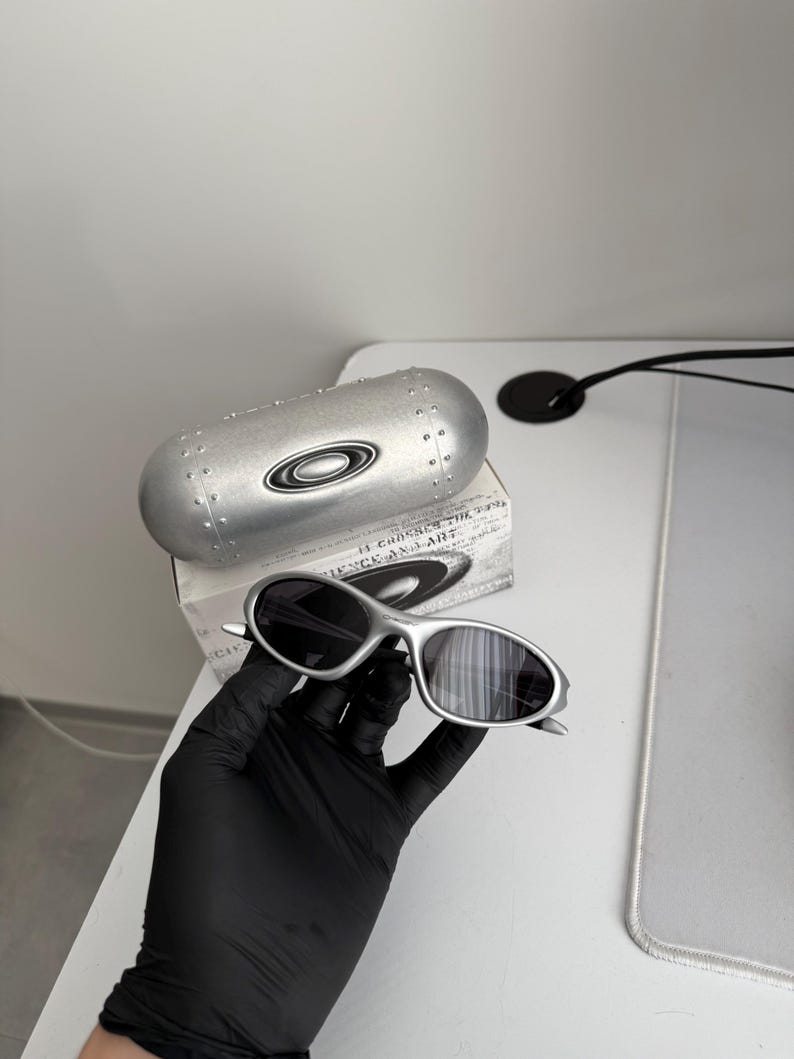 Puede incluir: Gafas de sol plateadas con lentes oscuras, presentadas con un estuche plateado a juego y una caja. Las gafas tienen un dise&ntilde;o elegante y moderno. El estuche presenta el logotipo de la marca.