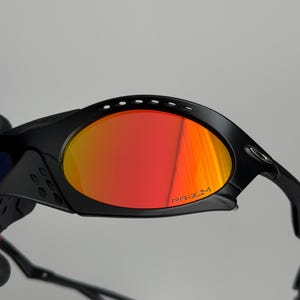 May include: Black sunglasses with red and orange lenses. The lenses have the word "PRIZM" printed on them. The sunglasses have a sporty design with a curved frame and arms.