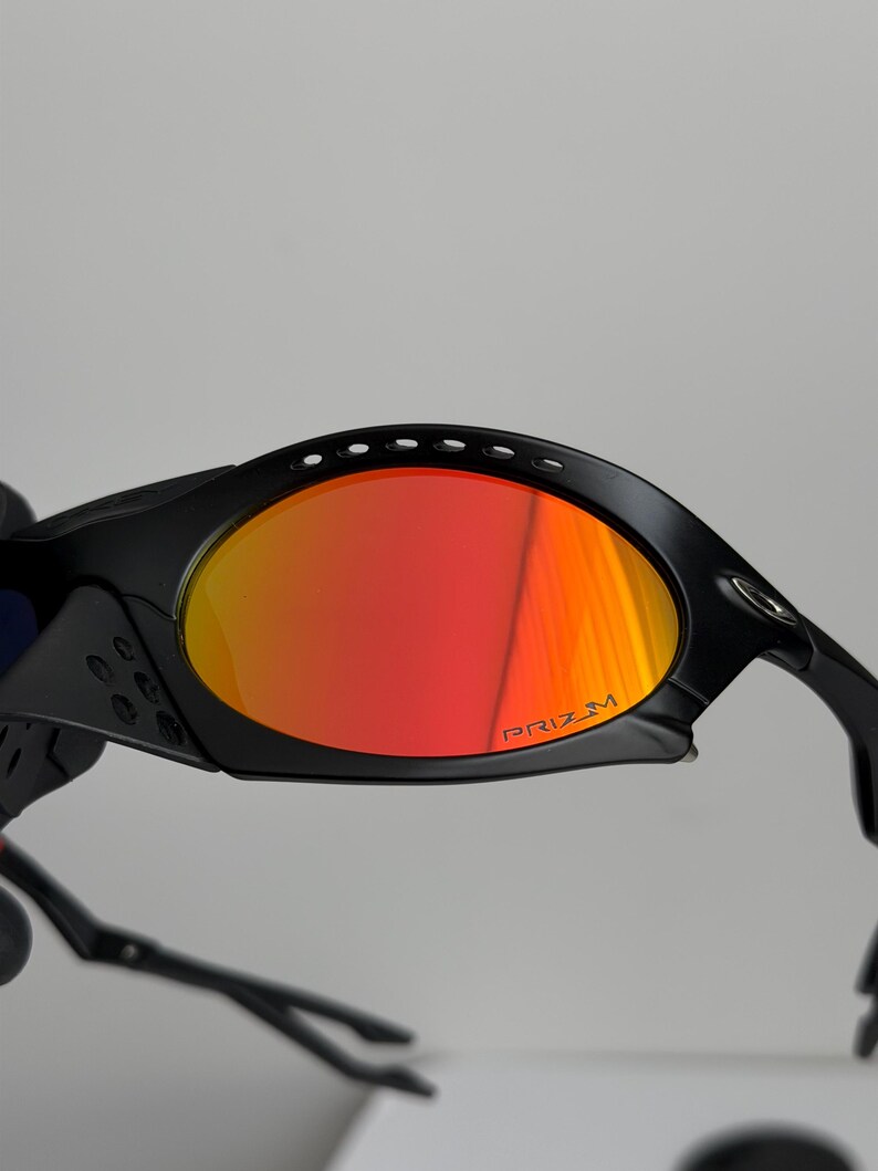 May include: Black sunglasses with red and orange lenses. The lenses have the word "PRIZM" printed on them. The sunglasses have a sporty design with a curved frame and arms.