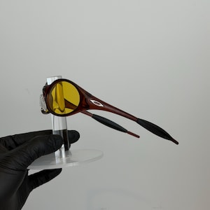 K&ouml;nnte beinhalten: Braune und gelb get&ouml;nte Sonnenbrille mit einem schlanken Design. Die Fassungen sind dunkelbraun und die Gl&auml;ser gelb. Die B&uuml;gel sind braun mit schwarzen Spitzen. Die Sonnenbrille wird auf einem transparenten St&auml;nder pr&auml;sentiert.