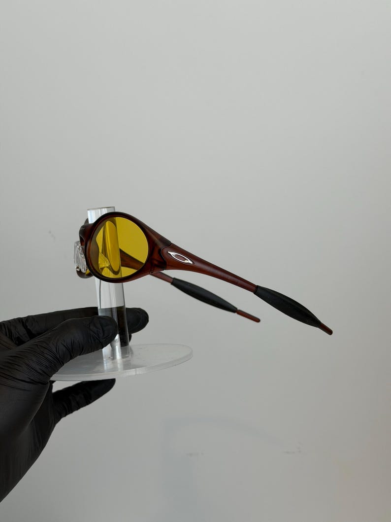 K&ouml;nnte beinhalten: Braune und gelb get&ouml;nte Sonnenbrille mit einem schlanken Design. Die Fassungen sind dunkelbraun und die Gl&auml;ser gelb. Die B&uuml;gel sind braun mit schwarzen Spitzen. Die Sonnenbrille wird auf einem transparenten St&auml;nder pr&auml;sentiert.