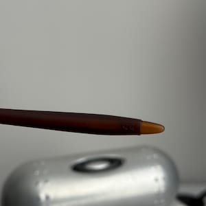May include: Close-up of a brown and amber-colored eyeglass arm. The arm is slender and has a rounded tip. The background features a silver case with the brand name.