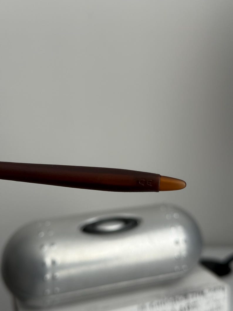 May include: Close-up of a brown and amber-colored eyeglass arm. The arm is slender and has a rounded tip. The background features a silver case with the brand name.