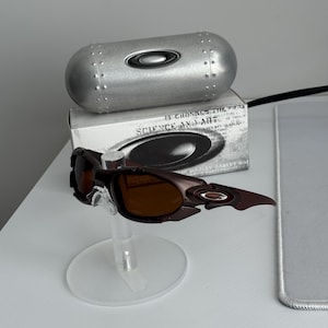 May include: Brown and black Oakley sunglasses with a matching case and box. The sunglasses have a unique, angular design with brown tinted lenses. The case is silver and cylindrical, with the Oakley logo.