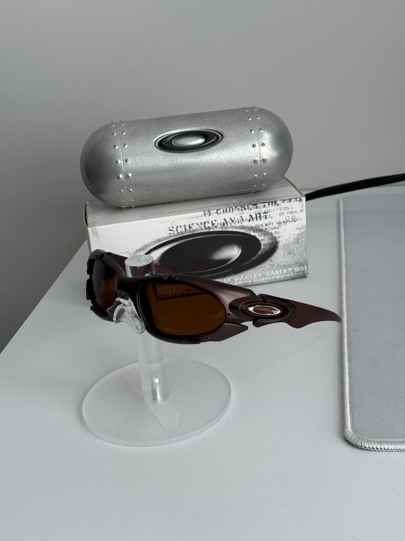 May include: Brown and black Oakley sunglasses with a matching case and box. The sunglasses have a unique, angular design with brown tinted lenses. The case is silver and cylindrical, with the Oakley logo.