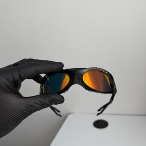 May include: Black sports sunglasses with orange and yellow mirrored lenses. The sunglasses have a sleek, aerodynamic design with a black frame and arms. The lenses reflect a gradient of colors.