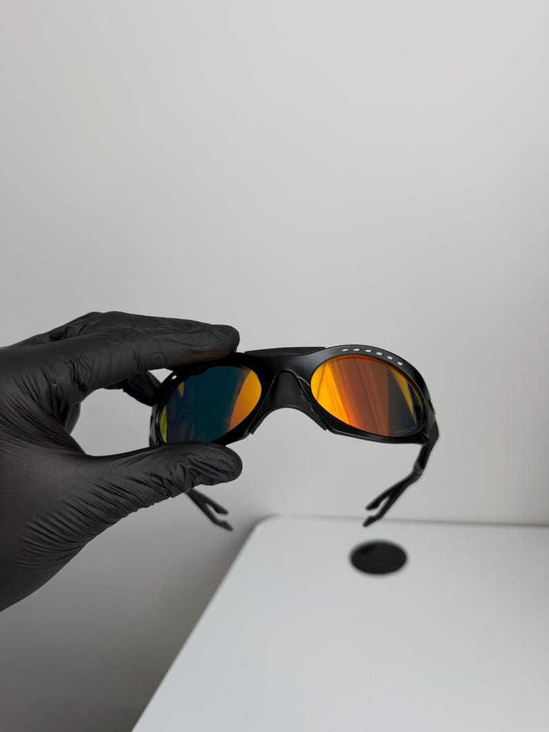 May include: Black sports sunglasses with orange and yellow mirrored lenses. The sunglasses have a sleek, aerodynamic design with a black frame and arms. The lenses reflect a gradient of colors.