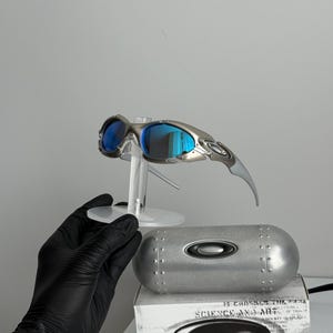 May include: Silver-framed sunglasses with blue mirrored lenses are displayed on a clear stand. A silver case and a white box with the text "SCIENCE AND ART" are below the sunglasses. The sunglasses have a sleek, modern design.