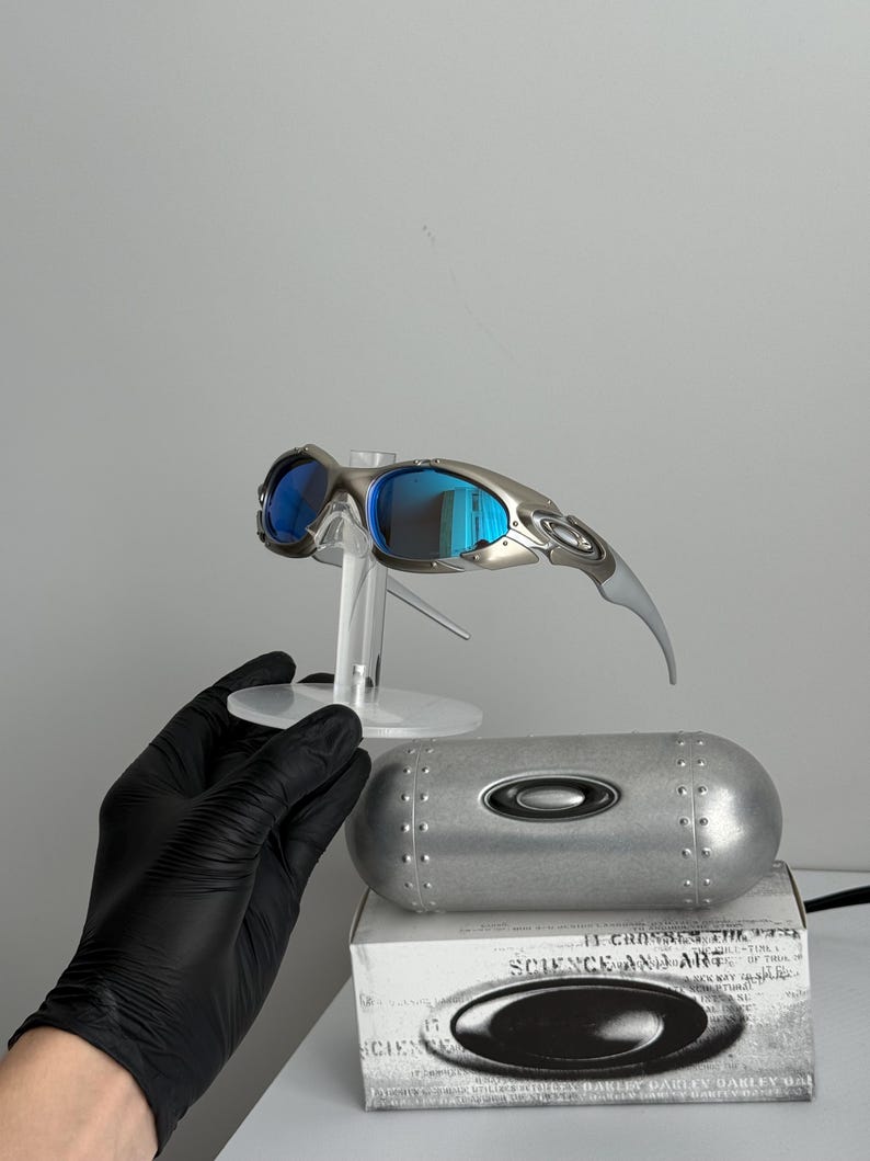 May include: Silver-framed sunglasses with blue mirrored lenses are displayed on a clear stand. A silver case and a white box with the text "SCIENCE AND ART" are below the sunglasses. The sunglasses have a sleek, modern design.