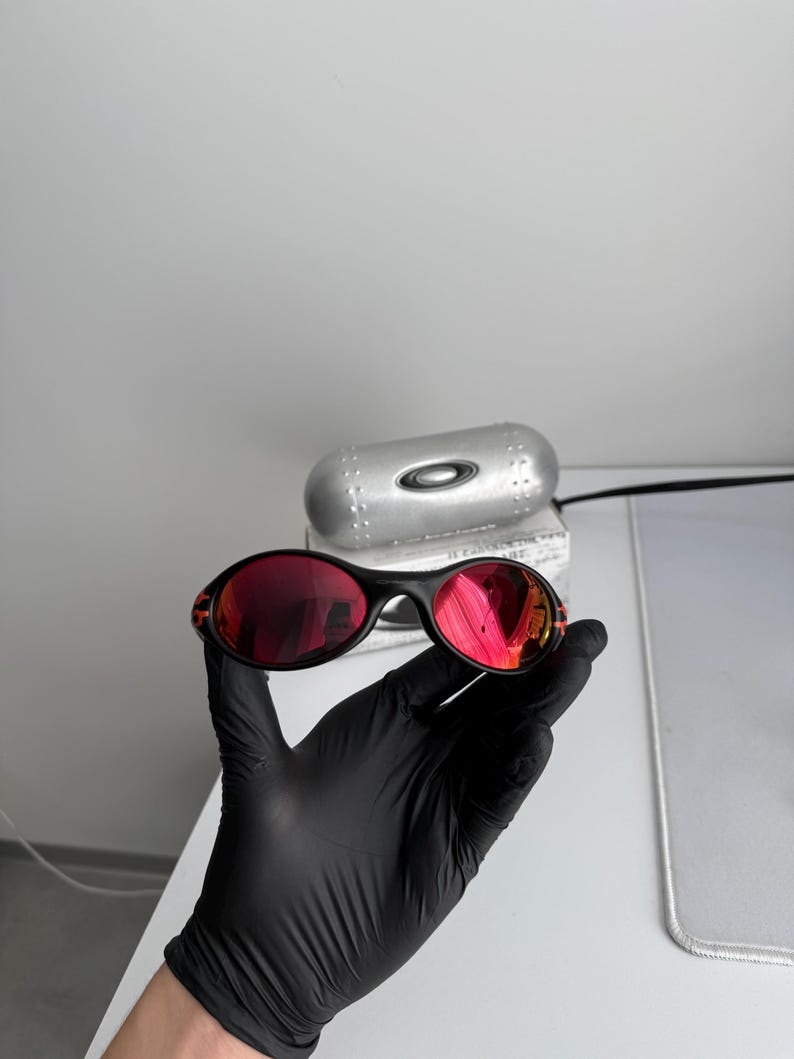 May include: Black sunglasses with red lenses are held by a gloved hand. The sunglasses have a sleek, oval shape and a black frame. The lenses reflect the surrounding environment. A silver case is in the background.