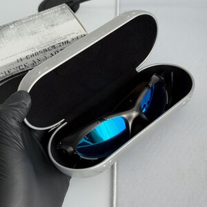 May include: Black sunglasses with blue reflective lenses rest inside an open silver case with a black interior. The case is rectangular with rounded edges and a riveted design. A box with text is partially visible.