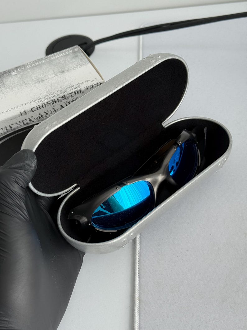 May include: Black sunglasses with blue reflective lenses rest inside an open silver case with a black interior. The case is rectangular with rounded edges and a riveted design. A box with text is partially visible.