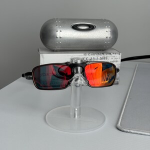May include: Black and brown sunglasses with red mirrored lenses are displayed on a clear stand. A silver case with the brand logo is behind the sunglasses. The lenses reflect a bright orange hue.