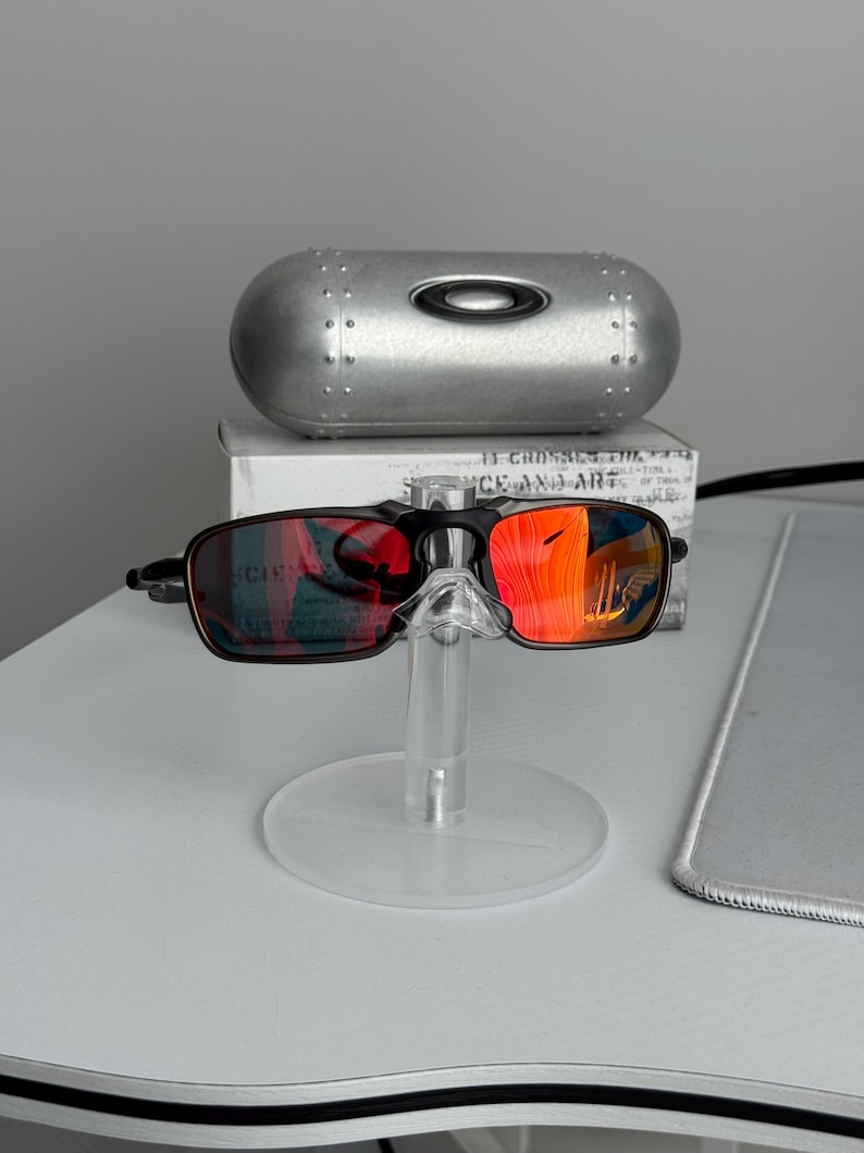 May include: Black and brown sunglasses with red mirrored lenses are displayed on a clear stand. A silver case with the brand logo is behind the sunglasses. The lenses reflect a bright orange hue.