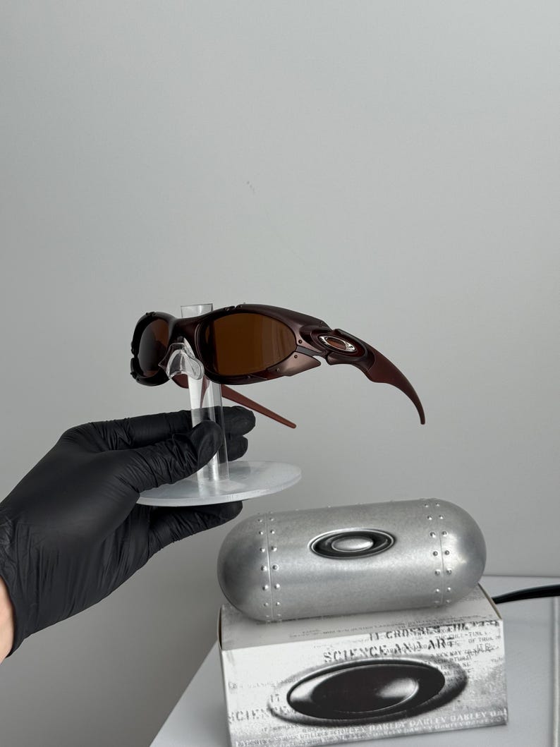May include: Brown Oakley sunglasses with a unique, angular design are displayed on a clear stand. A silver, cylindrical case and a box with the text "SCIENCE AND ART" are also visible. The sunglasses have brown lenses and a dark brown frame.