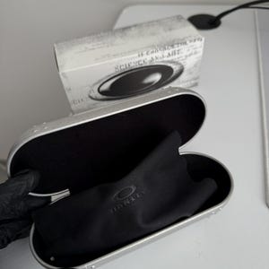 May include: Open silver glasses case with a black interior and a black microfiber cloth. The cloth has the brand name "OAKLEY" printed on it. A white box with the text "SCIENCE AND ART" is in the background.