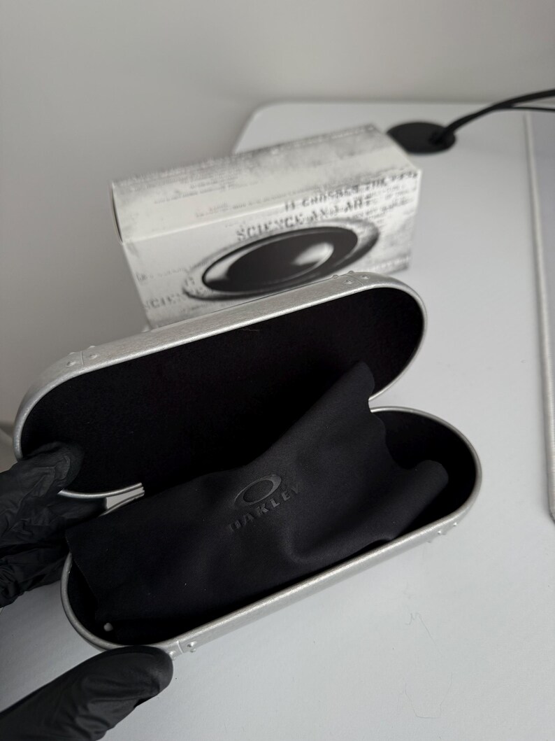 May include: Open silver glasses case with a black interior and a black microfiber cloth. The cloth has the brand name "OAKLEY" printed on it. A white box with the text "SCIENCE AND ART" is in the background.