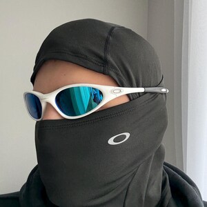 May include: White-framed sunglasses with blue mirrored lenses. The sunglasses are worn with a black balaclava. The Oakley logo is visible on the balaclava. The sunglasses have a sporty, modern design.