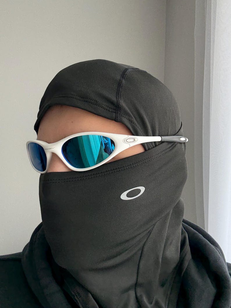 May include: White-framed sunglasses with blue mirrored lenses. The sunglasses are worn with a black balaclava. The Oakley logo is visible on the balaclava. The sunglasses have a sporty, modern design.