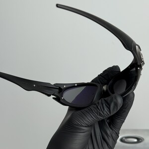 May include: Black sunglasses with dark lenses. The sunglasses have a sleek, modern design with a curved frame and arms. The lenses are dark, providing shade and protection from the sun. The sunglasses are held by a gloved hand.