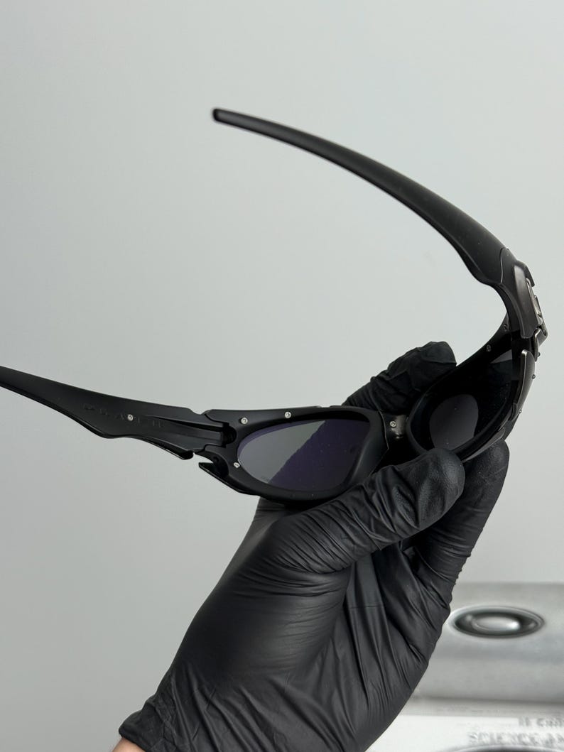 May include: Black sunglasses with dark lenses. The sunglasses have a sleek, modern design with a curved frame and arms. The lenses are dark, providing shade and protection from the sun. The sunglasses are held by a gloved hand.