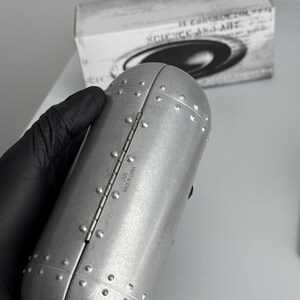 Pu&ograve; includere: Custodia in metallo argentato rivettata con un design a cerniera. La custodia &egrave; cilindrica con una parte superiore e inferiore arrotondate. Il testo "CASE MADE IN CHINA" &egrave; visibile. Una scatola con la scritta "SCIENCE AND ART" &egrave; sullo sfondo.
