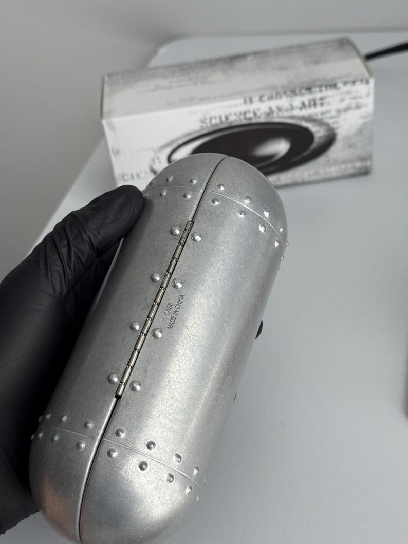 Pu&ograve; includere: Custodia in metallo argentato rivettata con un design a cerniera. La custodia &egrave; cilindrica con una parte superiore e inferiore arrotondate. Il testo "CASE MADE IN CHINA" &egrave; visibile. Una scatola con la scritta "SCIENCE AND ART" &egrave; sullo sfondo.