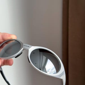 May include: Silver sunglasses with dark lenses. The sunglasses have a sleek, modern design with a curved frame and black temple tips. The lenses reflect the surrounding environment.