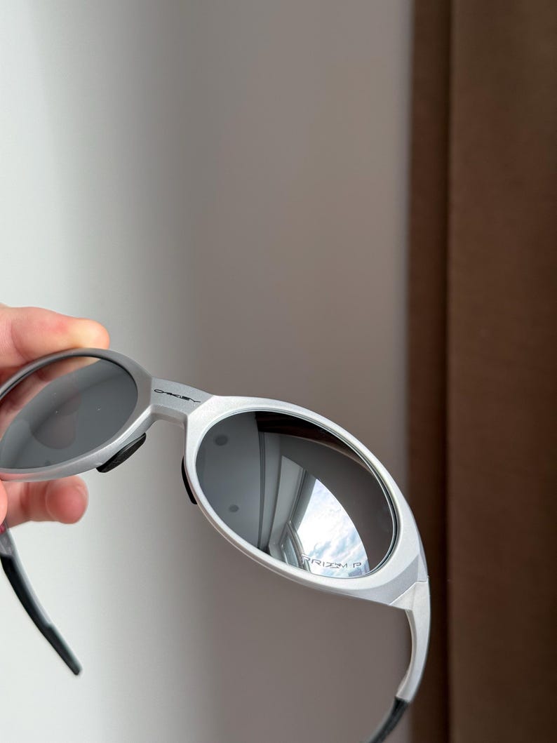 May include: Silver sunglasses with dark lenses. The sunglasses have a sleek, modern design with a curved frame and black temple tips. The lenses reflect the surrounding environment.