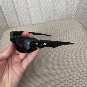 May include: Black sunglasses with dark lenses. The sunglasses have a sleek, modern design with a curved frame and arms. The Oakley logo is visible on the side of the frame.