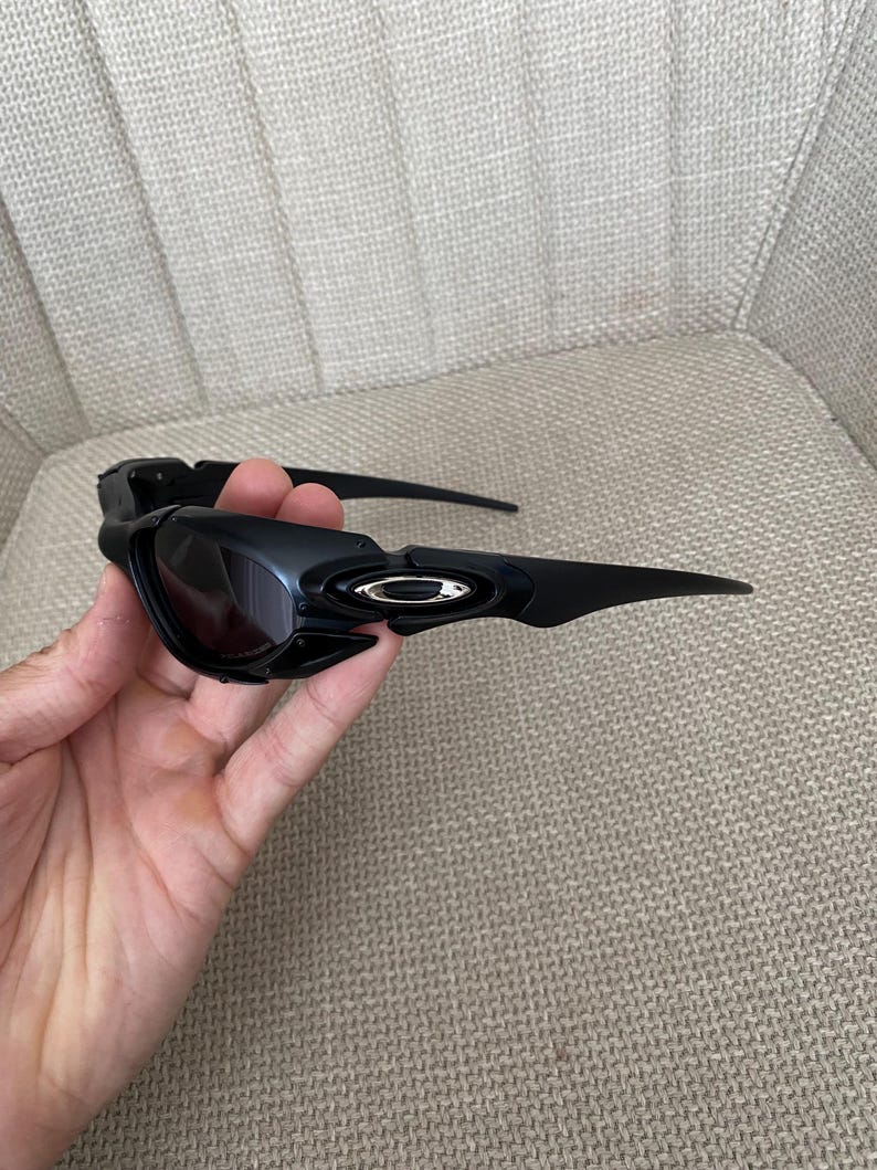 May include: Black sunglasses with dark lenses. The sunglasses have a sleek, modern design with a curved frame and arms. The Oakley logo is visible on the side of the frame.