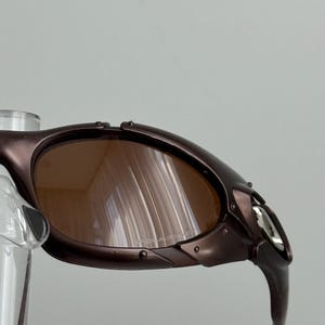 May include: Close-up of brown sunglasses with a metallic frame. The lenses are a warm brown color, and the frame has a dark, almost purple hue. The sunglasses have a sporty, wraparound design.