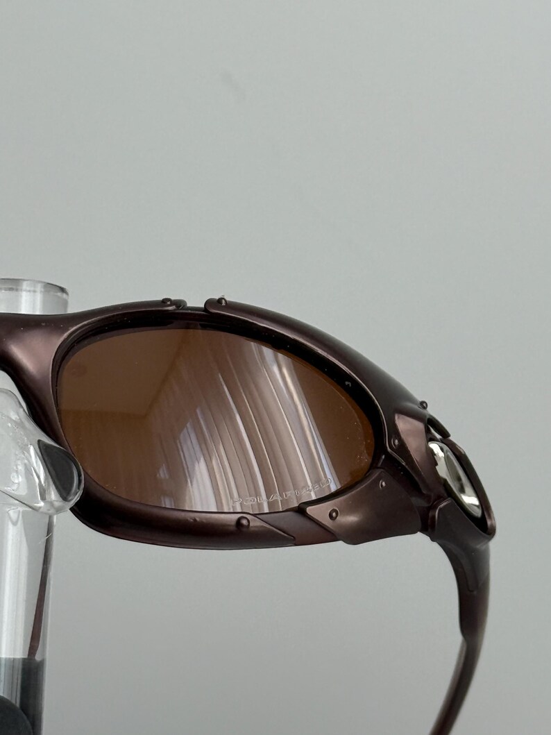 May include: Close-up of brown sunglasses with a metallic frame. The lenses are a warm brown color, and the frame has a dark, almost purple hue. The sunglasses have a sporty, wraparound design.