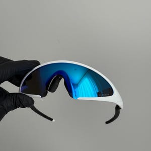 Puede incluir: Gafas de sol deportivas blancas con lentes azules espejadas. Las gafas tienen un dise&ntilde;o curvo y aerodin&aacute;mico con patillas negras y montura blanca. Las lentes reflejan un tono azul brillante, y la montura tiene la palabra "PRIZM".