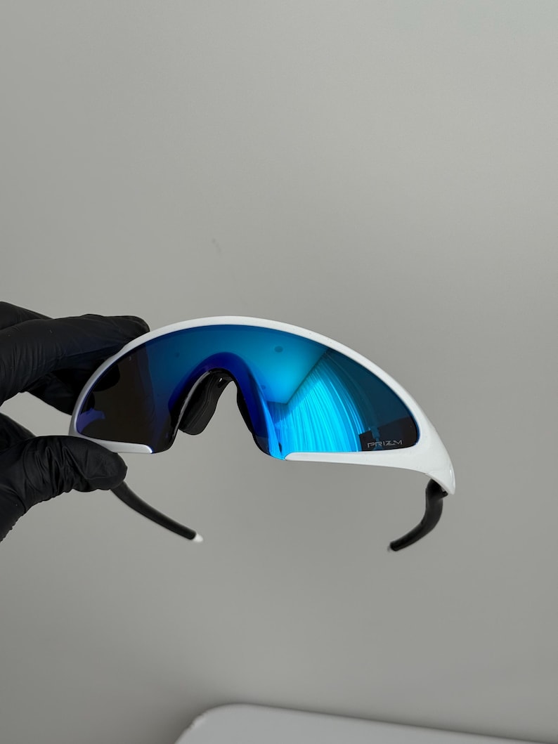 Puede incluir: Gafas de sol deportivas blancas con lentes azules espejadas. Las gafas tienen un dise&ntilde;o curvo y aerodin&aacute;mico con patillas negras y montura blanca. Las lentes reflejan un tono azul brillante, y la montura tiene la palabra "PRIZM".