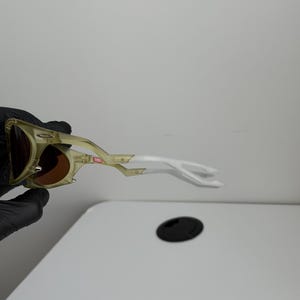May include: Sunglasses with a gold and white frame and brown lenses. The design is distinctive, with a curved shape and a small red accent. The frame is made of a lightweight material.