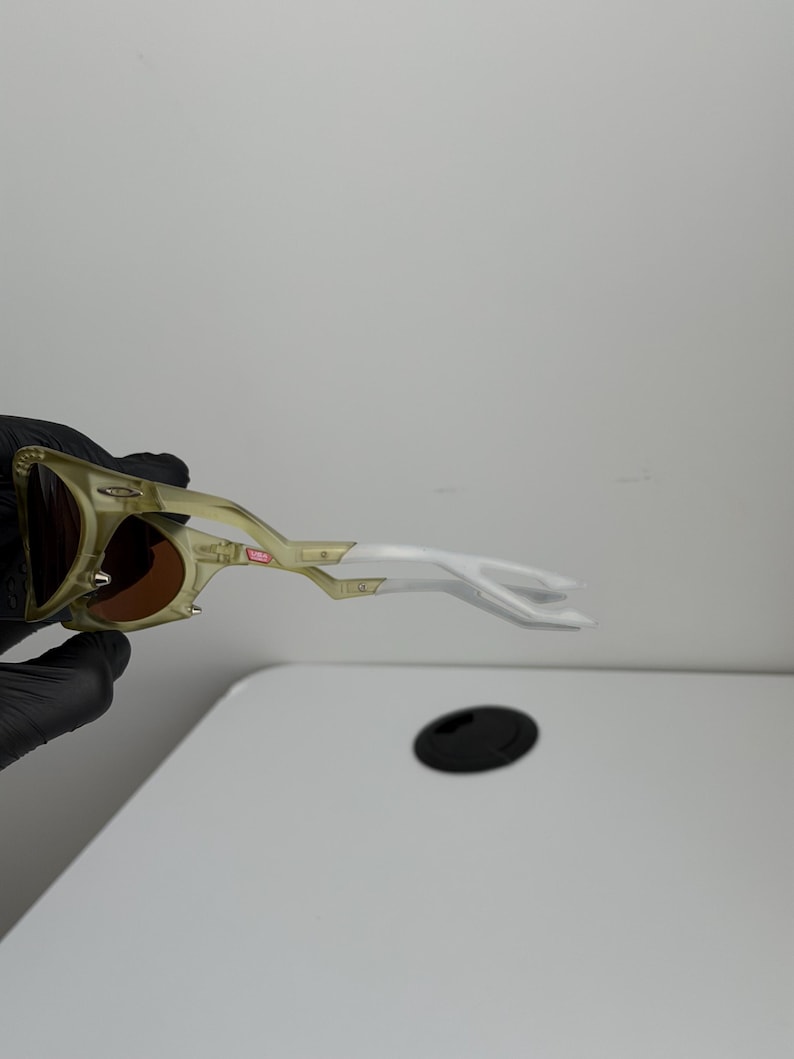 May include: Sunglasses with a gold and white frame and brown lenses. The design is distinctive, with a curved shape and a small red accent. The frame is made of a lightweight material.