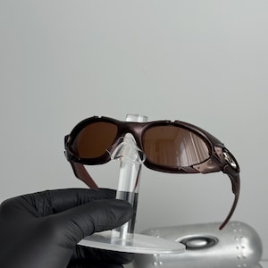 May include: Brown-tinted sunglasses with a dark brown frame. The sunglasses are displayed on a clear stand. The image shows the sunglasses from a side angle, highlighting the curved design of the frame and lenses.