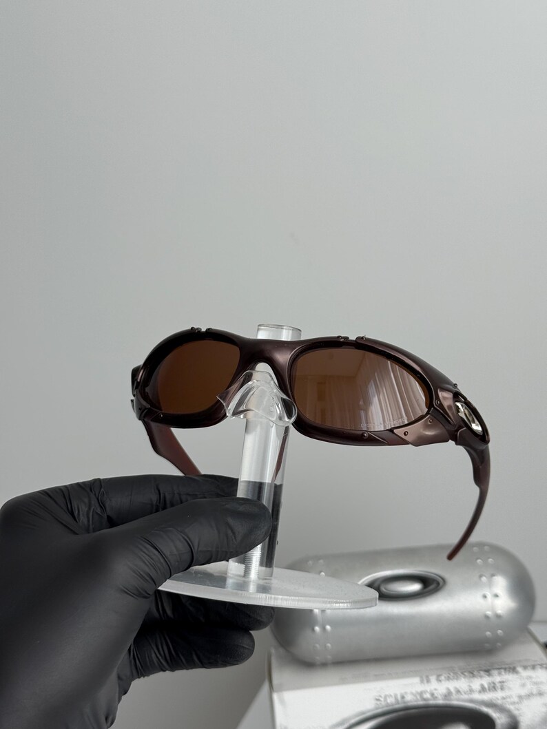 May include: Brown-tinted sunglasses with a dark brown frame. The sunglasses are displayed on a clear stand. The image shows the sunglasses from a side angle, highlighting the curved design of the frame and lenses.