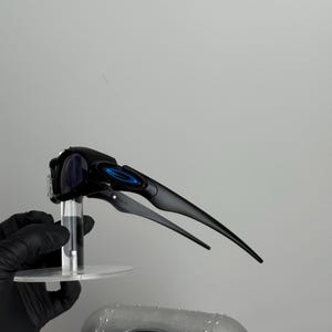 May include: Black sports sunglasses with blue accents and a sleek, aerodynamic design. The sunglasses have a curved frame and dark lenses. The arms extend back with a streamlined shape. The sunglasses are held up by a clear stand.