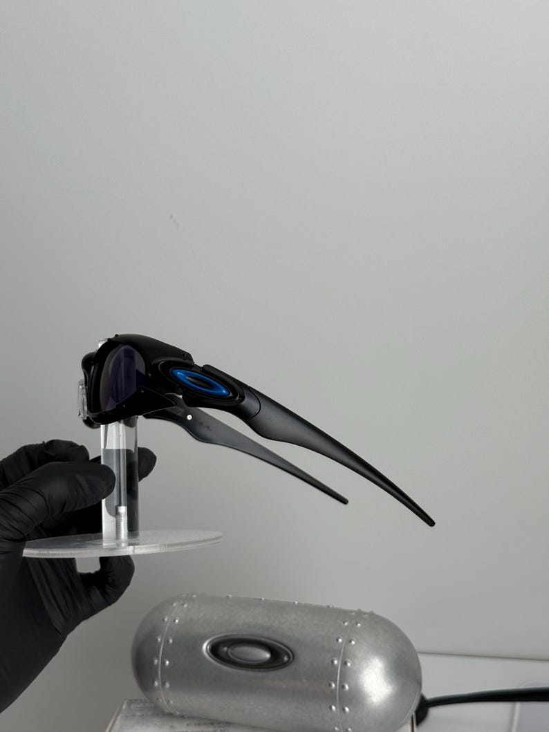 May include: Black sports sunglasses with blue accents and a sleek, aerodynamic design. The sunglasses have a curved frame and dark lenses. The arms extend back with a streamlined shape. The sunglasses are held up by a clear stand.