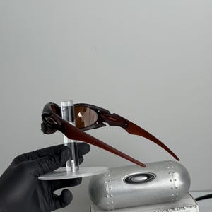 May include: Brown and black sunglasses with a unique frame design. The sunglasses are displayed on a clear stand, with a silver case and a box with the text "SCIENCE AND ART" visible in the background.