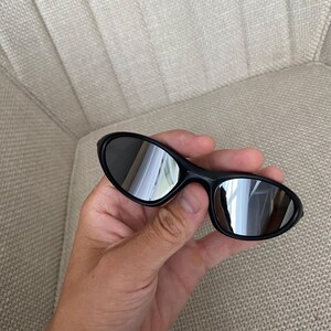Puede incluir: Gafas de sol negras con lentes reflectantes. Las gafas tienen forma ovalada y se sostienen en una mano. Las lentes reflejan el entorno.