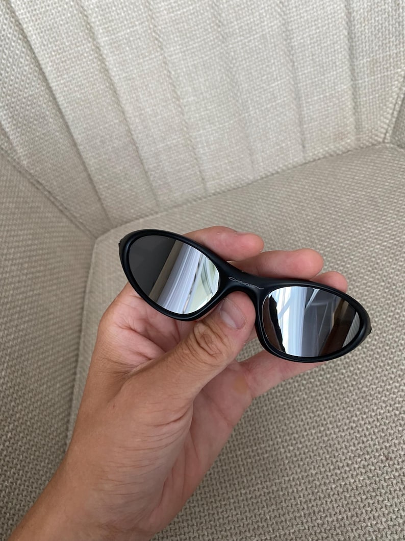 Puede incluir: Gafas de sol negras con lentes reflectantes. Las gafas tienen forma ovalada y se sostienen en una mano. Las lentes reflejan el entorno.