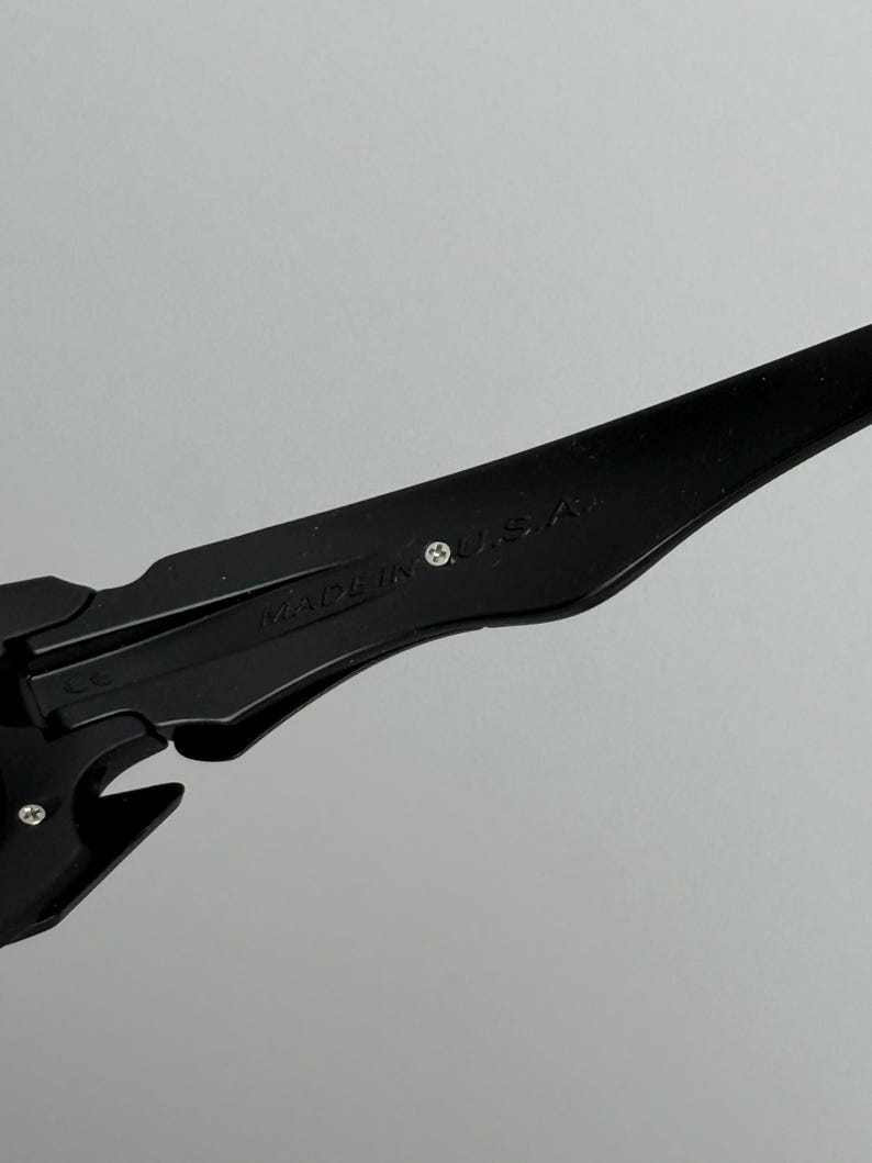May include: Close-up of black sunglasses with the text "MADE IN U.S.A." printed on the arm. The sunglasses have a sleek, modern design with a matte finish. The frame is a solid black color.