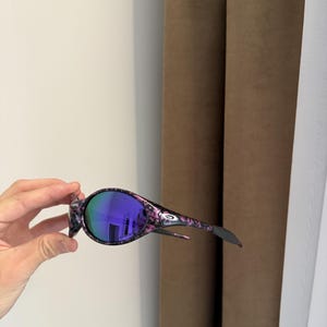 May include: Oval-shaped sunglasses with a black and pink floral pattern frame and purple lenses. The sunglasses have a sleek, modern design with a curved shape and a small logo on the temple.