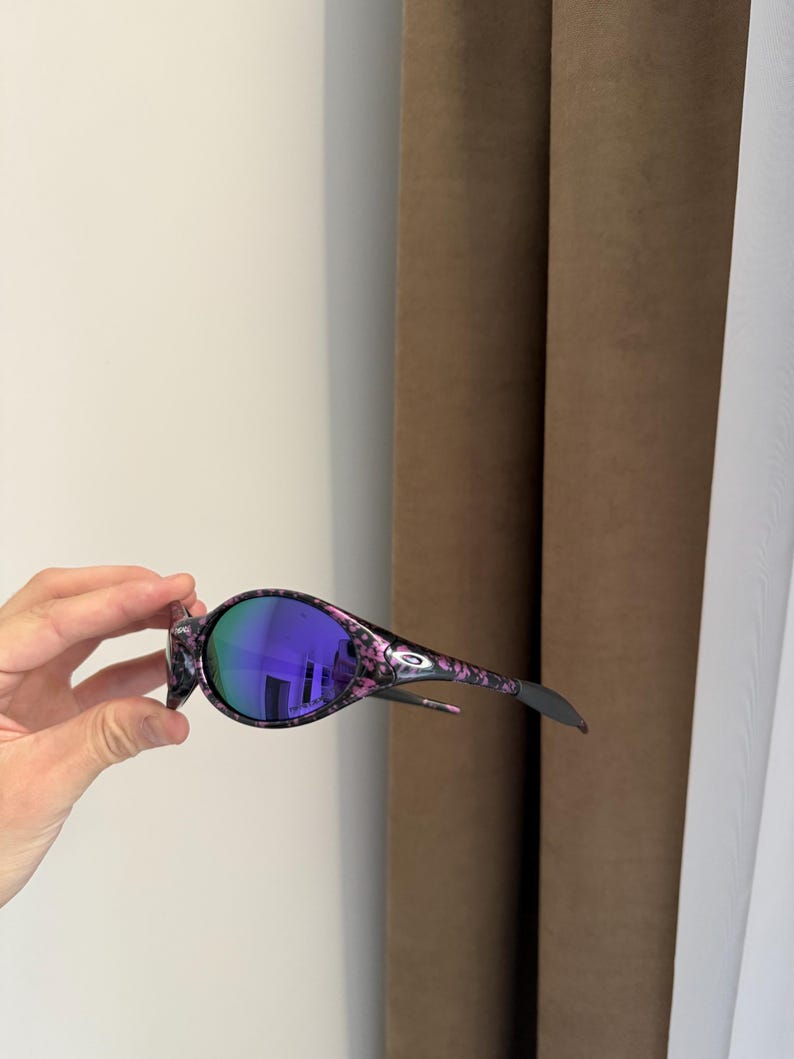 May include: Oval-shaped sunglasses with a black and pink floral pattern frame and purple lenses. The sunglasses have a sleek, modern design with a curved shape and a small logo on the temple.